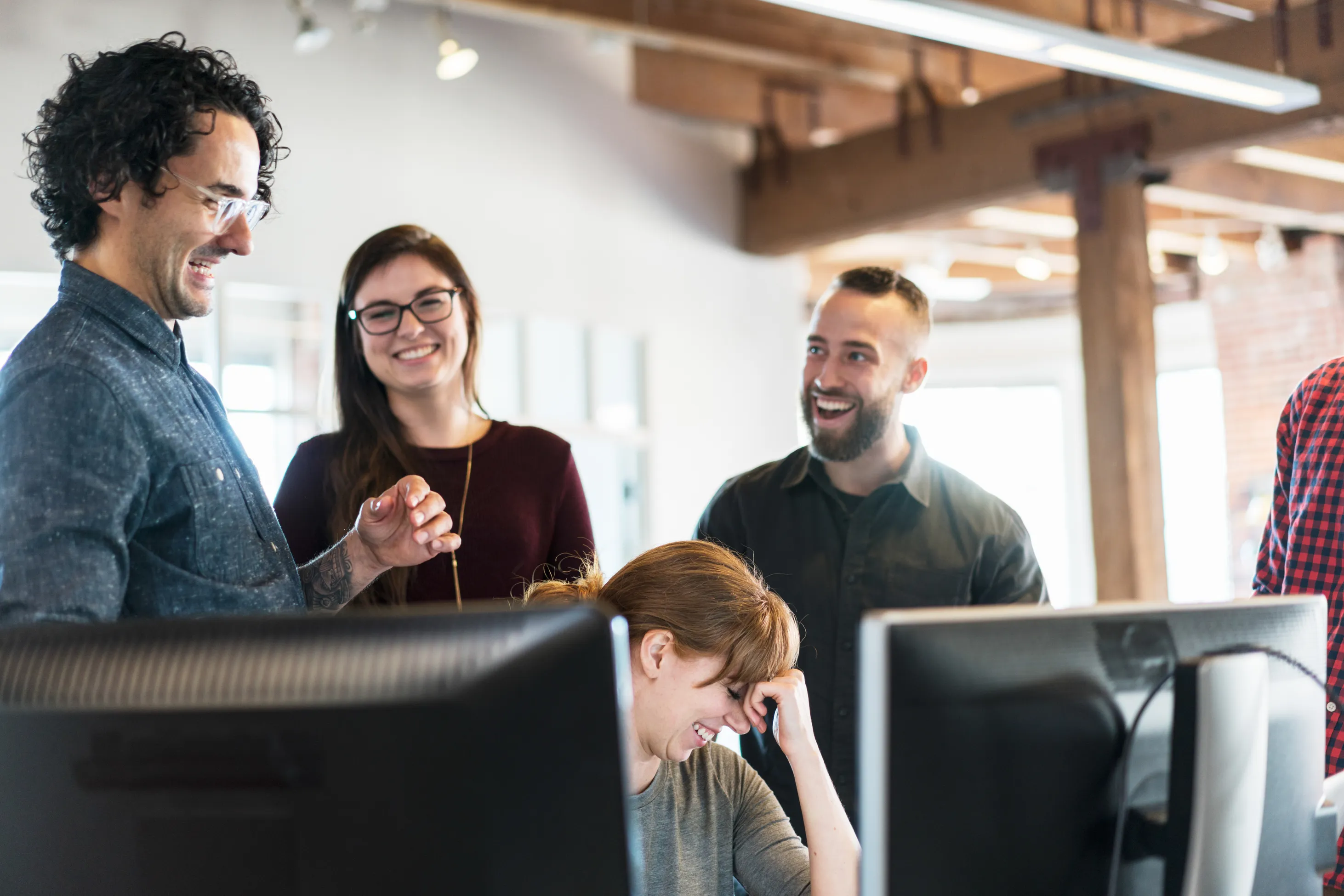 Four professionals in an office space laughing at a desk with computer monitors.