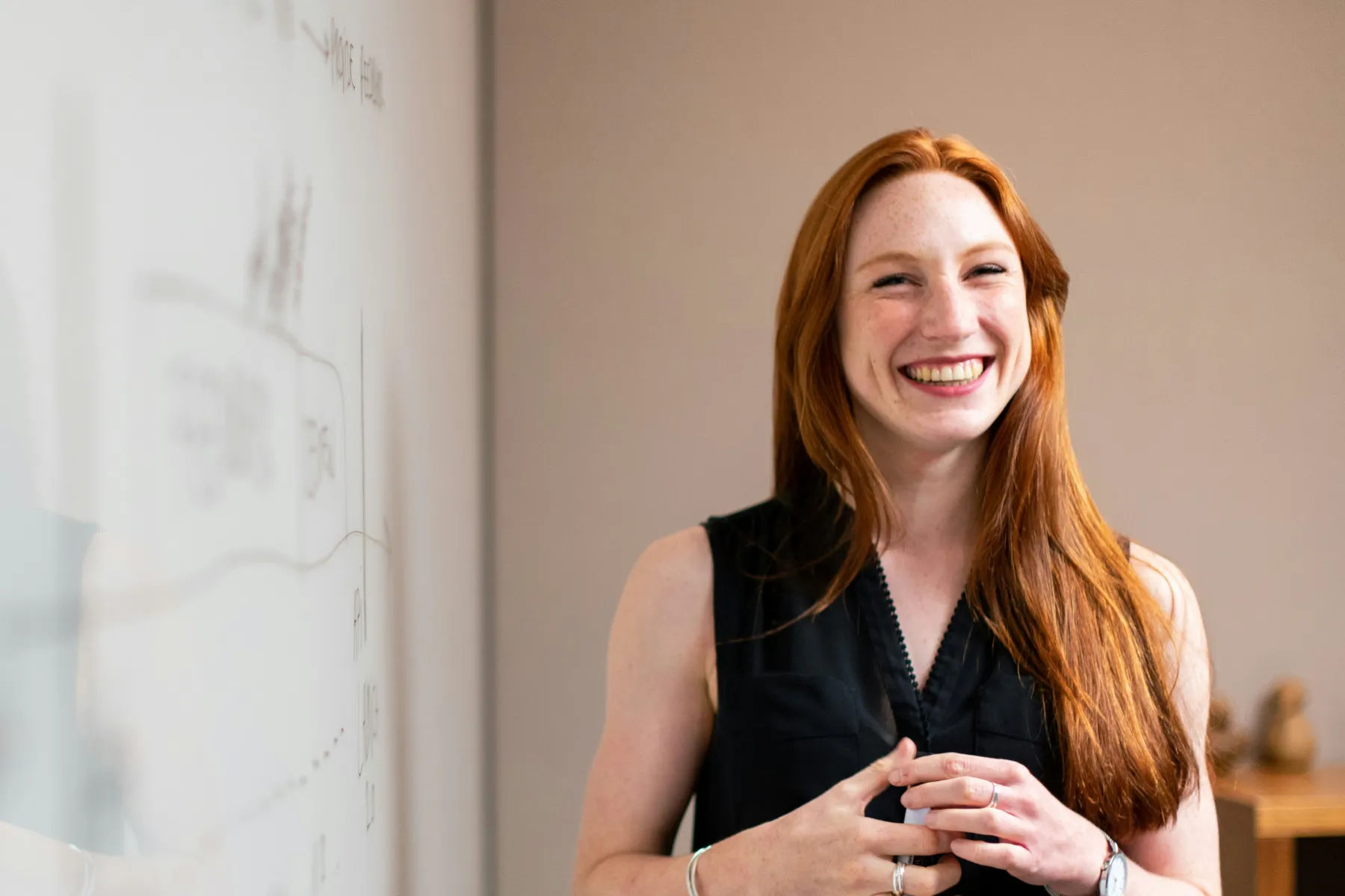 A person with long red hair smiles while standing next to a whiteboard with writing on it. The person is wearing a sleeveless black top.