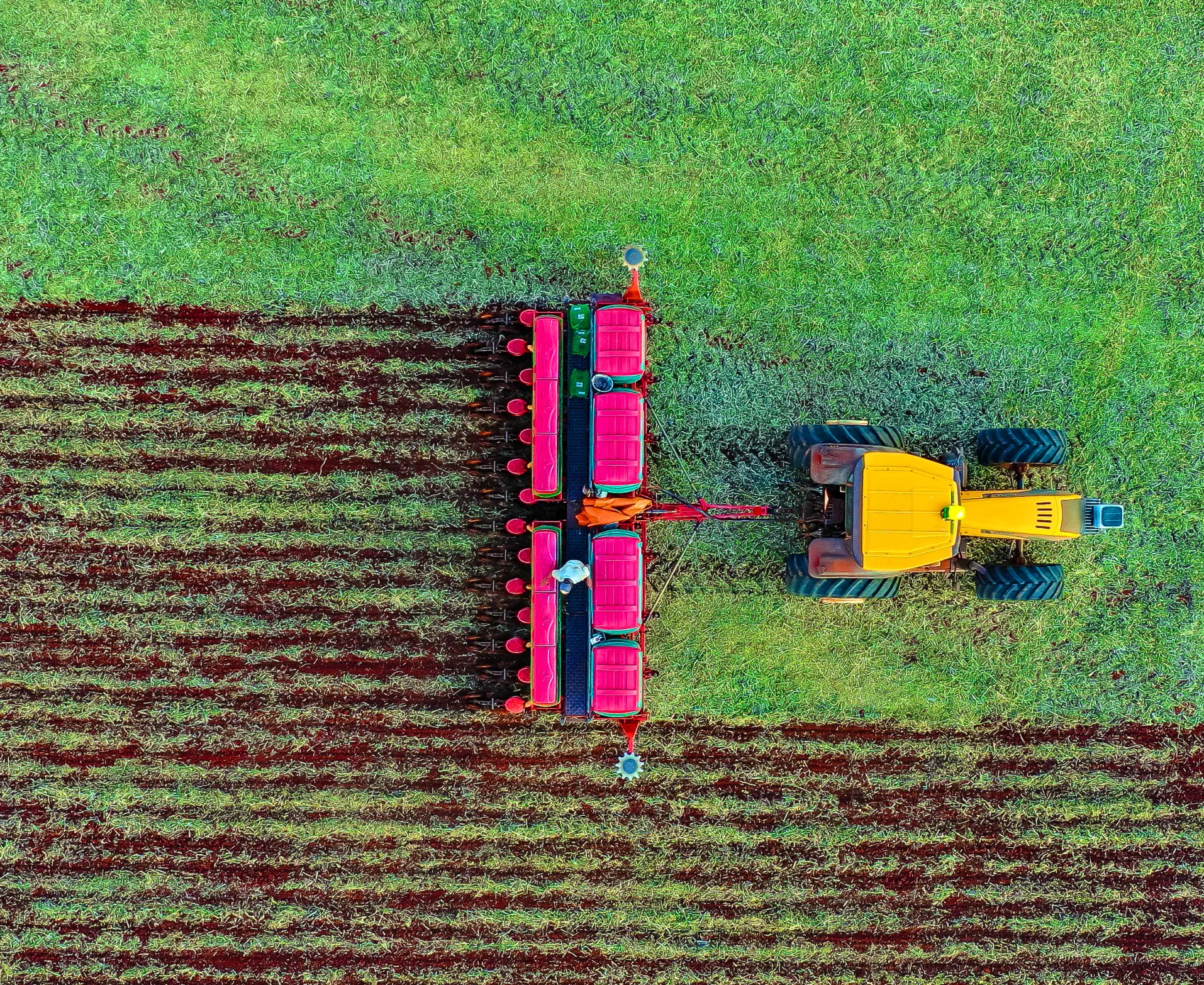 Aerial view of a bright yellow tractor with large tires pulling a red and green plowing attachment on a grassy field. The plow has created evenly spaced rows of freshly tilled soil, contrasting with the untouched green grass.